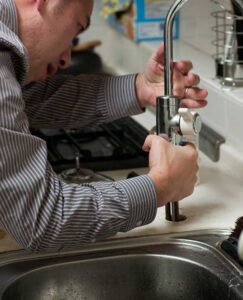 The image shows a man in a striped, long-sleeved shirt working on installing or repairing a kitchen faucet. He is using his hands to adjust or secure the faucet, which is of a modern, cylindrical design. The man is leaning over a stainless steel sink, and the background includes a blurred view of a typical kitchen, with a dish rack and stove visible. His focus and the use of manual tools suggest that he is performing a task commonly associated with a handyman or a plumber.