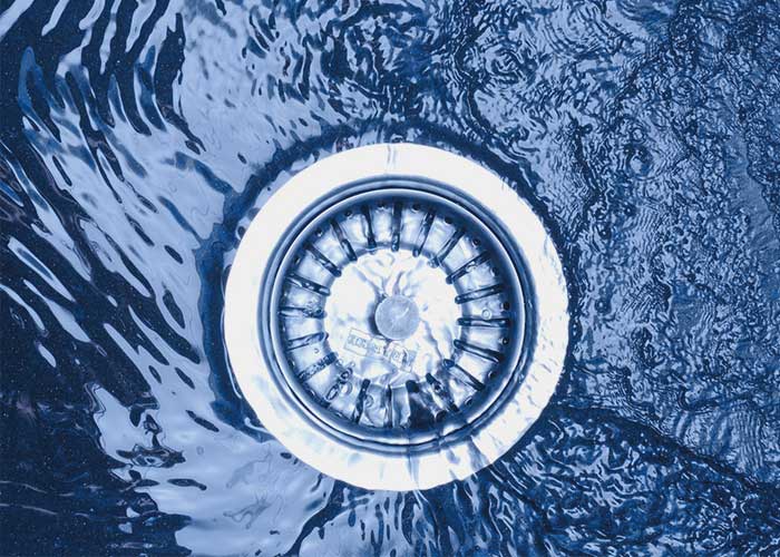 The image presents a close-up view of water swirling around a sink drain. The water's surface has ripples and the light reflecting off of it gives a metallic blue hue, emphasizing the fluid motion. The drain itself is in sharp focus, showing the typical design of a kitchen or bathroom sink drain with a central protective grid to prevent debris from entering the plumbing
