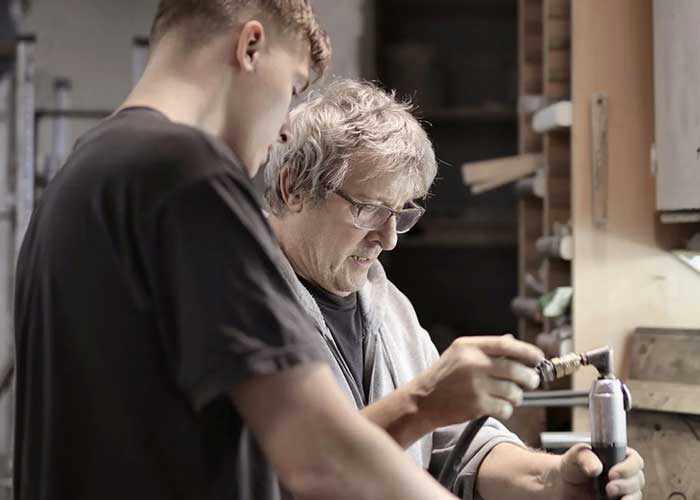 The image captures a moment of mentorship or apprenticeship in a woodworking or craft workshop. It shows an older man with grey hair and glasses, wearing a dust apron, teaching a younger individual who is attentively observing. They are both focused on a task at hand, where the older man appears to be guiding the younger one on how to use a hand tool, possibly a chisel or screwdriver, on a small object. The background suggests a workshop environment with tools and wooden materials. This scene represents the transfer of practical skills, likely related to handyman work or craftsmanship.