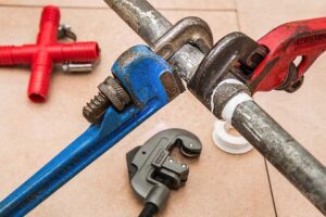 The image displays a close-up of two large wrenches, a blue one and a red one, in use on plumbing fixtures. The blue wrench is attached to a metallic pipe, while the red wrench is tightening or loosening a fitting on a white PVC pipe. In the background, out of focus, are additional red PVC connectors and what appears to be a black adjustable wrench. The tools and fittings are on a beige tiled floor, indicating a work area for plumbing repairs or installation. This setup is typical of a handyman's or plumber's work environment, showcasing the practical tools of the trade.