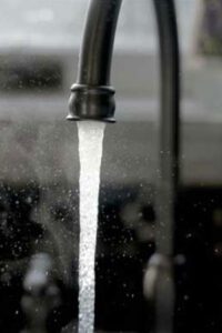 The image shows a close-up of a kitchen faucet with water flowing out steadily. The faucet appears to be modern in design, with a high arc and a black finish. The water stream is captured with a high shutter speed, freezing the motion and creating a crisp visual of the water droplets. The background is out of focus, which draws the eye to the faucet and the clear water stream in the foreground.