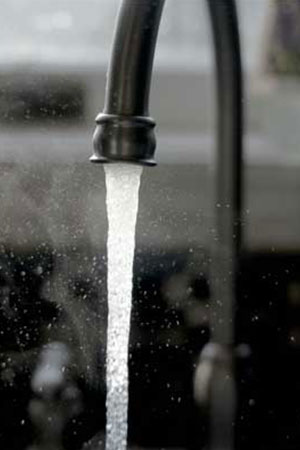 The image shows a close-up of a kitchen faucet with water flowing out steadily. The faucet appears to be modern in design, with a high arc and a black finish. The water stream is captured with a high shutter speed, freezing the motion and creating a crisp visual of the water droplets. The background is out of focus, which draws the eye to the faucet and the clear water stream in the foreground.
